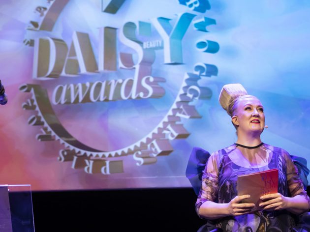 Dam med kub på huvudet står framför stor logotyp DAISY BEAUTY AWARDS